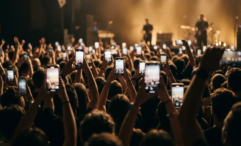 A concert crowd in the evening holding up glowing smartphones toward a warmly lit stage.