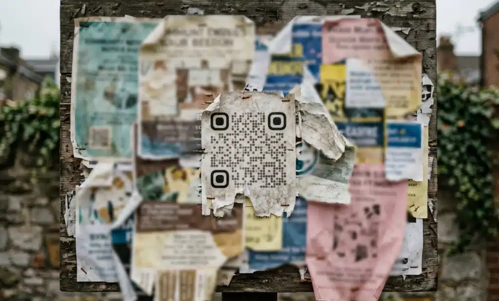 A torn and ignored QR code flyer among many overlapping posters on a cluttered noticeboard