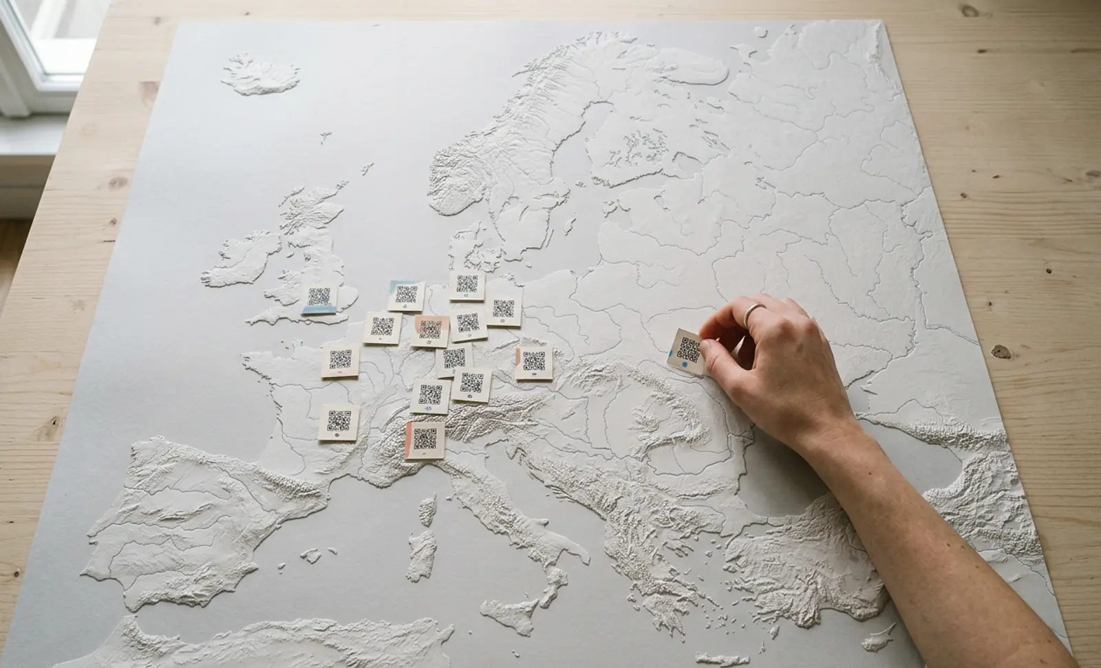 A hand placing printed QR code cards across a white relief map of Europe on a wooden table