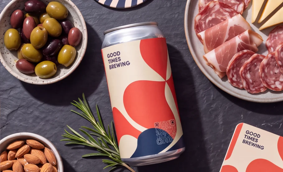 A beer can with a transparent QR code printed into its red and cream label, on a slate board with olives, almonds and charcuterie