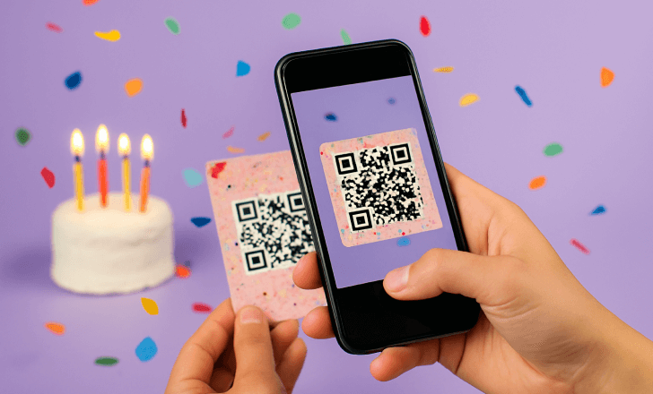 Close de uma pessoa escaneando um QR Code festivo em um cartão decorado, usando o celular, com um bolo de aniversário e confetes ao fundo.